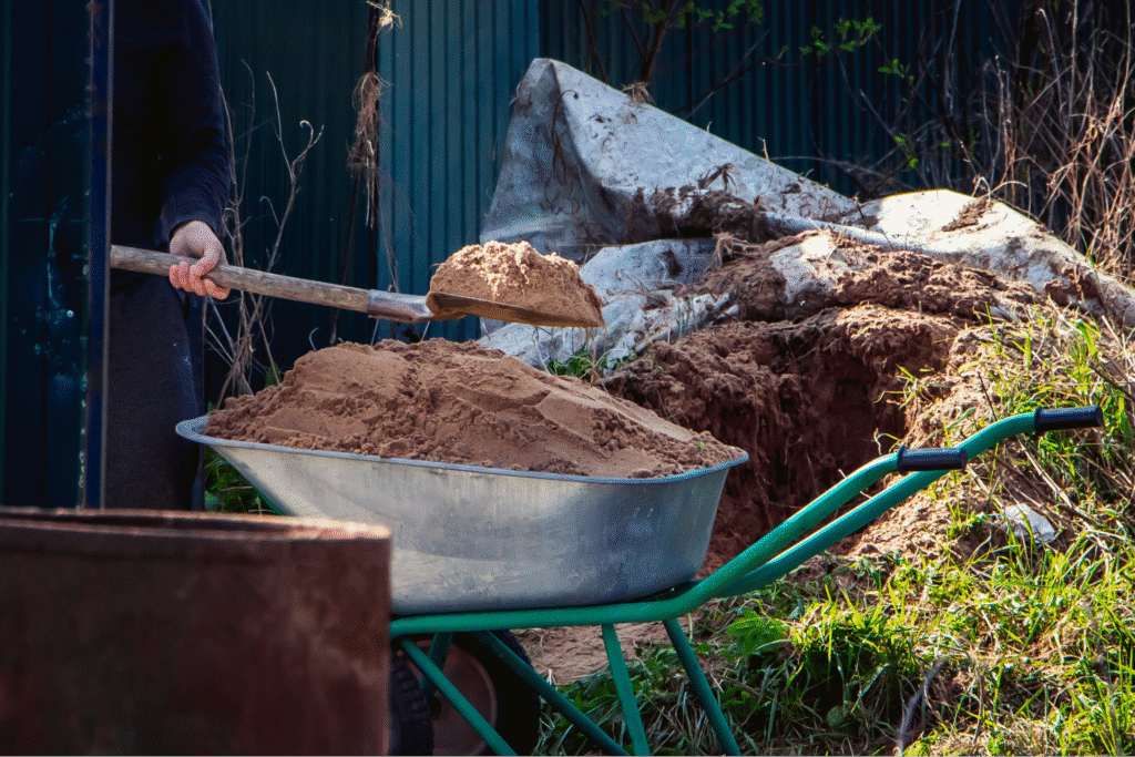 Person using a wheelbarrow to transport sand in a garden setting.