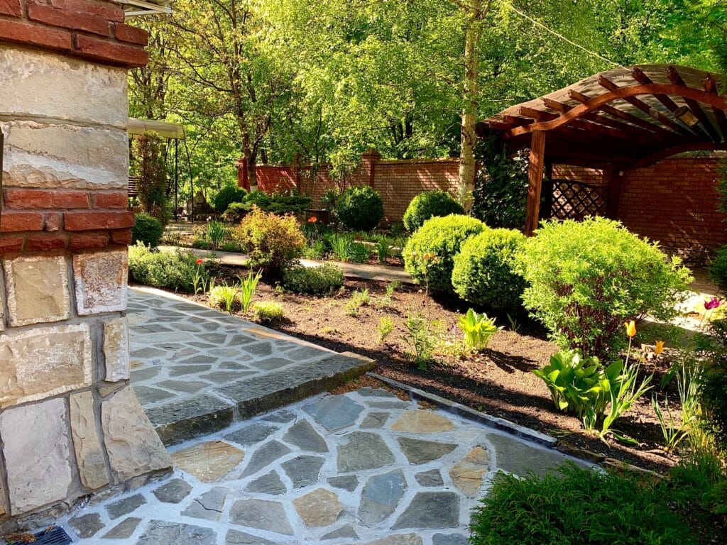 Lush garden path with flowering plants, stone walkway, and wooden gazebo.