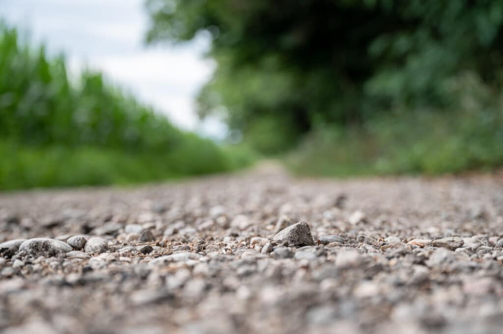 Gravel country road lined with lush green fields and pebbles in close-up view.