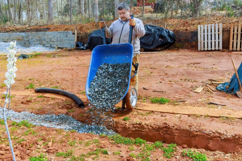 Man using a wheelbarrow to spread gravel in a construction area.