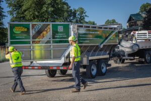 Workers guide a trailer filled with agricultural material on a sunny day.