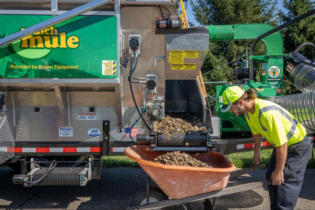 Landscaping crew member operates a mulch mule machine, loading organic material into a wheelbarrow.