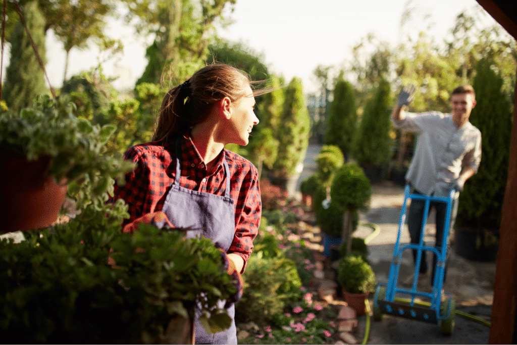 Working in a vibrant garden, a woman admires plants while a man waves nearby.