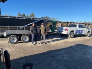 Two workers stand beside a truck and trailer on a sunny day.