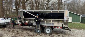 Man standing by trailer-mounted equipment in a wooded area, showcasing professional tools.