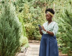 Woman in a garden using a smartphone among lush greenery and trees.