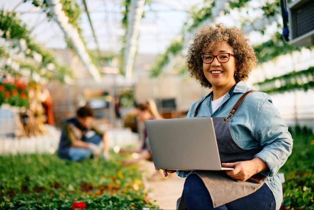Smiling woman in greenhouse uses laptop for AI in landscaping planning