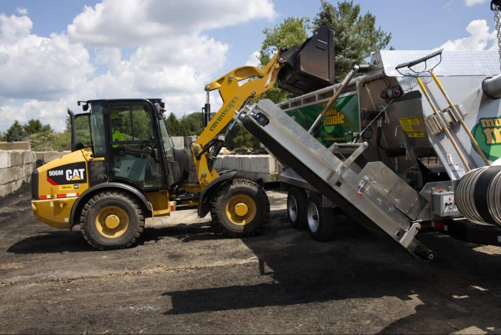 Caterpillar loader unloading materials into a Mulch Mule at a mulch yard under a blue sky.