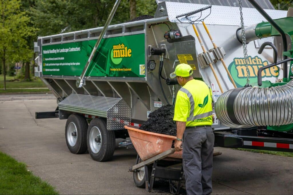 Worker loading mulch into a wheelbarrow from a green landscaping truck.